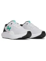 Championes de running Under Armour modelo Charged Surge 4, color blanco con detalles en negro y logo en verde agua. Presentan parte superior de malla transpirable con superposiciones sintéticas, plantilla Deluxe Comfort System y entresuela Charged Cushioning para mayor amortiguación y respuesta.