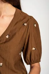 Blusa de viscosa color marrón, con escote en V, mangas cortas abullonadas y cierre frontal con botones. Cuenta con pequeños detalles bordados distribuidos por la prenda.