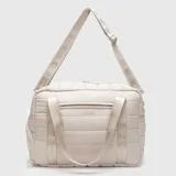 Bolso deportivo tipo duffle color beige, con diseño acolchado y acabado mate. Cuenta con doble asa de mano, correa ajustable para el hombro con logo grabado, cierre principal superior, bolsillo frontal con cierre y compartimento lateral independiente para calzado.
