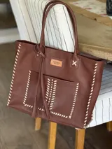 Bolso tipo tote de cuero color marrón con costuras blancas en contraste, bolsillo externo, doble asa de hombro y cierre superior con cremallera.