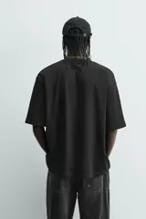 Camiseta negra de corte relaxed fit, cuello redondo y manga corta. Presenta un estampado frontal en blanco con efecto spray que simula dos figuras volando, y texto en un óvalo que dice "SUCCESS CHASER".