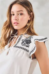 Blusa de color blanco con detalles bordados en negro, cuello redondo con cordones ajustables y mangas cortas con volados.