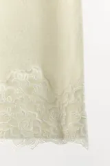 Falda midi de tejido ligero color crema o beige muy claro, con cintura elástica de color beige más oscuro. El bajo está adornado con una aplicación de encaje floral a tono.