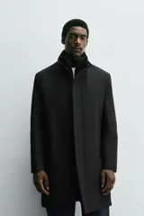 Abrigo negro de corte recto con cuello subido y doble cuello desmontable mediante cremallera interior. Cuenta con manga larga, bolsillos de vivo en la cadera, bolsillo interior, abertura central en la espalda y cierre frontal de cremallera oculta por solapa con botones a presión.