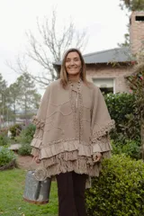 Ruana de punto color beige con textura, cuello alto con volados y lazo regulable. Presenta detalles de volados aplicados en lino en mangas y ruedo, con terminación de flecos artesanales en la parte inferior.