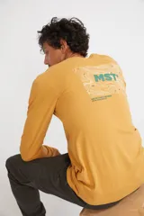 Remera de manga larga color mostaza, con cuello redondo y estampado frontal con las siglas MST en contorno verde.