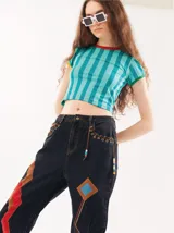 Pantalón de jean tiro alto tipo mom-fit con parches de colores en las piernas, apliques de tiento de cuero trenzado con canutillos alrededor de la cintura y costuras decorativas en zigzag.