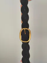 Cinturón trenzado de estilo artesanal confeccionado en material símil gamuza color negro, con hebilla ovalada metálica en tono dorado.