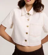 Camisa cropped de manga corta confeccionada en sarga de algodón, color crudo. Presenta cuello clásico, cierre frontal con botones metálicos, un bolsillo aplicado en el pecho y costuras pespuntadas en contraste.