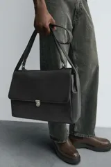 Bolso tipo bandolera color negro, con compartimento principal con cierre metálico y bolsillo interior con cremallera. Tiene un bolsillo liso en la parte trasera, asa de mano y asa de hombro ajustable.