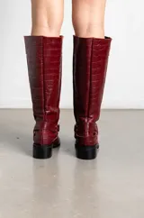 Botas de caña alta en cuero con textura de cocodrilo, color bordó. Presentan un diseño de punta cuadrada, taco bajo y una correa decorativa con argolla metálica y remaches en la zona del tobillo.
