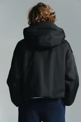 Anorak corto de color negro con acolchado interior, cuello subido y capucha. Presenta mangas largas con puños elásticos, bolsillos delanteros con cierre y bajo ajustable con cordones. Cierre frontal con cremallera termosellada y botones a presión.