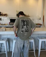 Remera oversize gris oscuro con corte holgado, mangas cortas y estampa trasera en blanco y negro con forma de estampilla que muestra a una persona con un gato y el texto 'Kaboa Industries'.