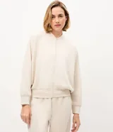 Campera bomber deportiva de mujer color beige, confeccionada en modal con cierre frontal por cremallera, cuello medio y bolsillos. Presenta mangas largas con puños elásticos y elástico en la cintura.