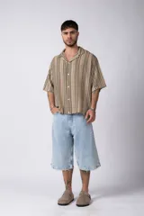 Camisa de manga corta con cuello solapa, corte holgado y cierre frontal de botones. Presenta un diseño de rayas verticales en tonos beige, negro y blanco.