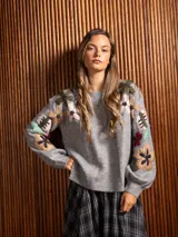 Buzo de cuello redondo en color gris, con mangas largas y diseño holgado. Presenta un bordado floral multicolor con detalles de cuentas en la parte frontal y en las mangas.