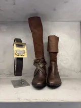 Bota de montar de caña alta, confeccionada en cuero marrón con combinación de texturas. Presenta un diseño con puntera redondeada, taco cuadrado y una correa decorativa cruzada con tachas metálicas en el tobillo.