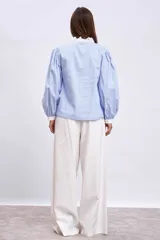 Blusa celeste con rayas verticales, cuello en V, mangas largas abullonadas con puños blancos y volantes verticales en el frente.