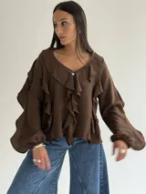 Blusa blanca de corte holgado con escote en V, decorada con volados frontales y en las mangas. Presenta mangas largas con puños elásticos y cierre frontal con botones.