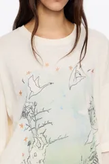 Camiseta oversize de algodón y poliéster color crema con cuello redondo y manga corta ancha. Incluye una manga interior ajustada de tul transparente con estampado sutil. Estampado frontal con diseño de paisaje, árboles, aves y texto.
