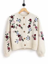 Cardigan de punto color marfil, con cuello redondo a la base, mangas largas y cierre frontal con botones. Presenta un delicado bordado artesanal de flores en tonos bordó, rosado y celeste con tallos y hojas verdes en el frente y las mangas.
