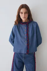 Camisa de algodón estilo denim con corte regular, cuello redondo y cierre frontal con botones. Presenta detalles de volados tejidos a mano en crochet en color azul marino con bordes rojos en el cuello y la cartera frontal.