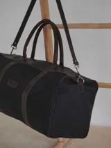 Bolso de viaje tipo duffel de lona negra con detalles en cuero tostado, asas de mano y correa ajustable para el hombro.