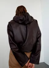 Chaqueta de cuero de napa marrón oscuro, de diseño oversize, con capucha ajustable, cierre frontal de cremallera, bolsillos laterales y bajo ajustable con cordón.