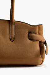 Cartera de hombro color tostado, con doble asa superior y detalle de tiras laterales ajustables. Cuenta con cierre magnético y etiqueta colgante.