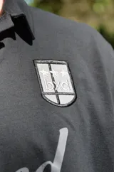 Chomba negra de corte cuadrado con cuello polo y mangas cortas. Presenta un escudo estampado en el pecho con diseño de fútbol retro y la palabra 'boxed' estampada en el frente.