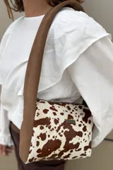 Cartera tipo bandolera con diseño de estampado animal print de vaca en tonos marrón y blanco. Presenta una correa ancha de gamuza color habano y una placa metálica con el logo de la marca en el frente.