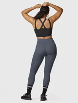 Calza deportiva de mujer color gris oscuro, con cintura alta y largo hasta el tobillo. Presenta un bolsillo lateral en la pierna derecha y un logo en la parte trasera de la cintura.