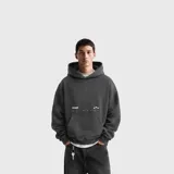 Hoodie de corte oversize en color gris oscuro con acabado lavado. Presenta capucha amplia, bolsillo frontal tipo canguro y un pequeño estampado gráfico en el pecho con estrellas y texto.