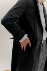 Tapado largo de jean azul oscuro, con cuello solapa y puños con detalle de cinta de terciopelo negro. Presenta costuras visibles que definen paneles y un logo bordado en la espalda.