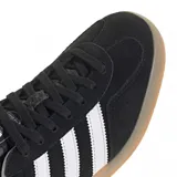 Championes Adidas Gazelle Indoor de diseño clásico, confeccionados en gamuza negra con las icónicas tres tiras laterales en blanco. Presentan el nombre del modelo en letras doradas, talón en contraste blanco y suela de goma color caramelo.