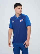 Remera polo deportiva Umbro del Club Nacional de Football, color azul con cuello en V y detalles en rojo y blanco. Incluye el logo de Umbro y el escudo del club.