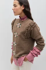 Sweater de lana color celeste, con cuello redondo, mangas largas y volados decorativos en los hombros. Presenta bordados florales artesanales en el frente y detalles de tejido calado vertical.