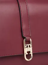 Cartera de hombro de cuero sintético color borgoña, con solapa frontal y cierre metálico con monograma TH. Posee correa ajustable y detalles metálicos dorados.