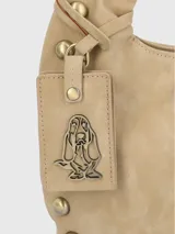 Cartera tipo hobo de Hush Puppies en color taupe, confeccionada en material con textura gamuzada. Presenta un diseño de media luna con tachas metálicas decorativas en los laterales y la base, asa única integrada y un dije colgante con el logo de la marca.