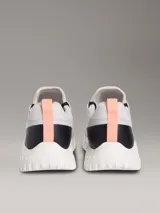 Zapatillas tipo runner Calvin Klein, de color gris con detalles en negro y coral. Presentan un diseño de malla perforada en la parte superior, cordones negros, suela chunky de EVA blanca y el logo de la marca en la lengüeta y el lateral.