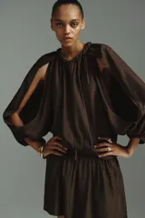 Vestido túnica corto de color marrón con leve brillo, corte oversized, cuello redondo con ribete trenzado y escote en V con tiras finas. Cuenta con mangas globo largas de corte raglán con abertura en los hombros, puños elásticos y cintura fruncida con vuelo en la falda.