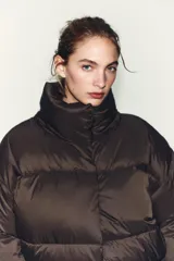 Campera puffer corta de color marrón oscuro, con diseño acolchado, cuello alto y capucha integrada. Presenta cierre frontal con botones magnéticos y mangas largas con puños elásticos.