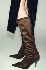 Bota alta de tacón fino, confeccionada en material con acabado de gamuza color marrón oscuro. Presenta una tira decorativa con hebilla metálica en la parte superior de la caña, cierre lateral mediante cremallera y punta fina.