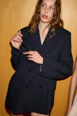 Blazer oversize de corte recto con cuello solapa y cierre frontal cruzado con botones. Presenta mangas largas con hombreras, trabillas en los hombros y grandes bolsillos de plastrón con solapa en el delantero. Incluye abertura en la espalda y forro interior.