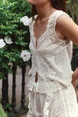 Top de algodón color crema con escote redondo, tirantes anchos y cierre frontal con botones. Presenta detalles de plisados, encaje combinado, bordados florales y aplicaciones de abalorios, con el bajo acabado en ondas.