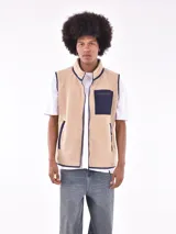 Chaleco de polar tipo corderito en color beige, con cuello alto y cierre frontal completo. Presenta un bolsillo de parche en el pecho en color azul marino con cierre de cremallera y ribetes en contraste en el mismo tono en sisas, cuello y bolsillos laterales.