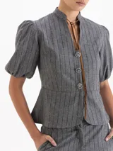 Chaqueta gris con diseño de rayas verticales y sutiles lentejuelas. Presenta cuello mao con lazo de gamuza, botones decorativos en forma de flor, recorte en la cintura y mangas abullonadas con puño elástico.