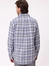 Camisa de manga larga con estampado de cuadros en tonos blanco, gris y azul. Presenta cuello clásico, cierre frontal con botones y un pequeño parche con logo en el pecho.