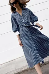 Vestido midi azul marino con rayas blancas, cuello solapa, escote en V y cierre frontal con botones. Cuenta con mangas abullonadas por debajo del codo con puños anchos a contraste, cinturón ajustable del mismo tejido con hebilla metálica y falda evasé.