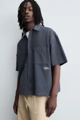 Camisa de algodón con estructura canvas, color rojo lavado, de corte relajado (relaxed fit) y manga corta. Posee cuello solapa, dos bolsillos de plastrón en el pecho y cierre frontal de botonadura. Presenta bordado y estampado de texto a contraste en el delantero.